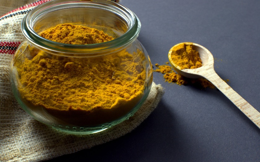 glass jar of orange coloured ground turmeric on a tea towel with a wooden spoon on the worktop next to it and turmeric on the spoon and counter