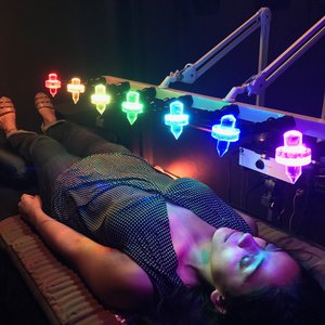woman lying on a mat with a metal arm holding seven crystals above her. The crystals are lit and coloured red, orange, yellow, green, teal, blue and pink and are positioned down the centre of the body in a line from head to toe with even spacing